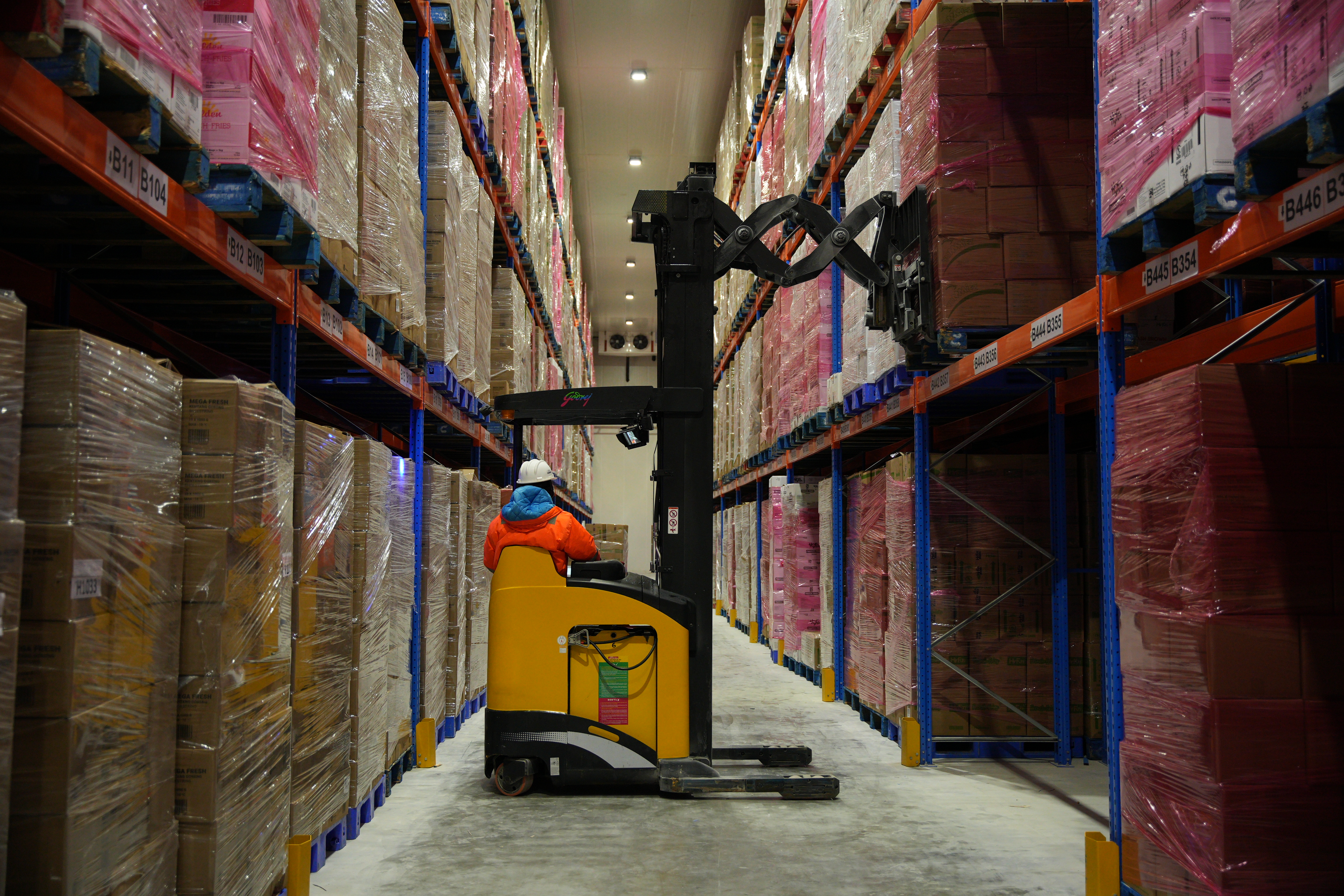 A worker in an orange jacket and hard hat operates a yellow forklift in Cold Chain Storage. Tall shelves stacked with goods create a symmetrical, organized scene.